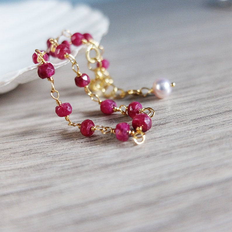 Op de afbeelding: Een delicate gouden kettingarmband met kleine, gefacetteerde robijn kralen en een enkele parel bedel.