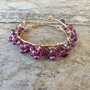 May include: A gold-colored wire bracelet adorned with clusters of deep purple, faceted beads. The bracelet features a double-band design with a clasp closure. The beads are a rich, dark color, adding a touch of elegance to the jewelry.