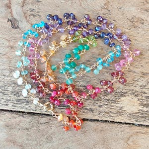 May include: A multi-colored gemstone necklace featuring faceted, teardrop-shaped beads in shades of red, orange, yellow, green, blue, and purple, all linked together with thin gold-toned wire and secured with a circular spring ring clasp.