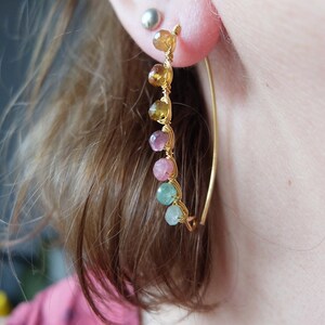 May include: Gold hoop earrings with a row of colorful gemstones. The gemstones are in shades of green, pink, and yellow.