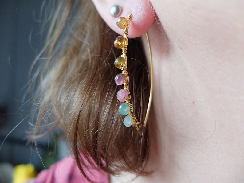 May include: Gold hoop earrings with a row of colorful gemstones. The gemstones are in shades of green, pink, and yellow.