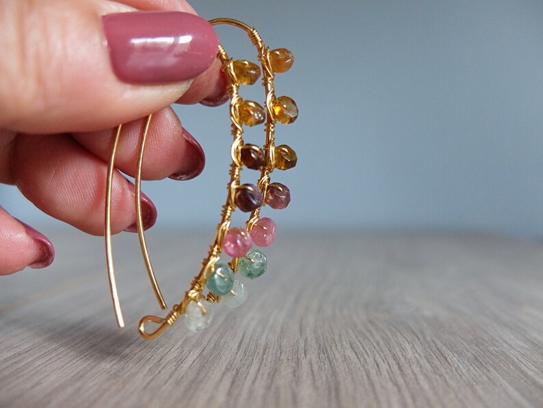 May include: Gold wire earrings with a cascade of colorful gemstones. The gemstones are in shades of green, pink, brown, and yellow.