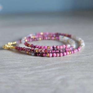 Puede incluir: Un brazalete de múltiples hilos con cuentas rosas, rojas y blancas. El brazalete está hecho de cuentas de piedras preciosas y cuentas doradas.