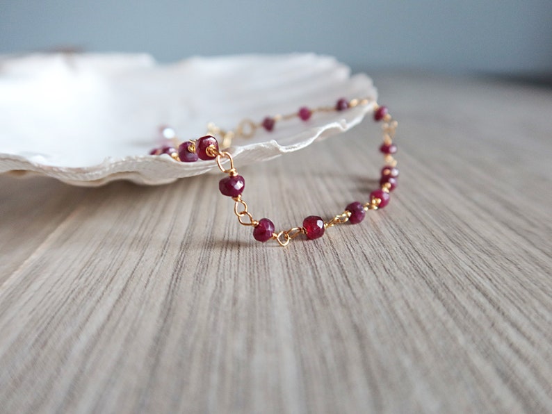 Op de afbeelding: Een delicate gouden kettingarmband met kleine, gefacetteerde robijnrode kralen.