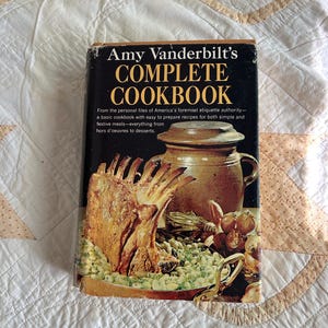 May include: A vintage cookbook titled "Amy Vanderbilt's Complete Cookbook" with a black cover and gold lettering. The cover features a food illustration of a roasted meat dish, a ceramic pot, and other food items. The book is on a white quilted surface.