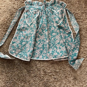May include: A teal apron with a floral pattern in white and gray. The apron has white trim and long ties. The fabric is a light cotton, and the apron is laying on a brown carpet.