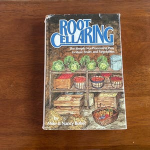 May include: A hardcover book titled Root Cellaring: The Simple No-Processing Way to Store Fruits and Vegetables by Mike & Nancy Bubel. The cover features a detailed illustration of a root cellar with shelves holding baskets of produce.