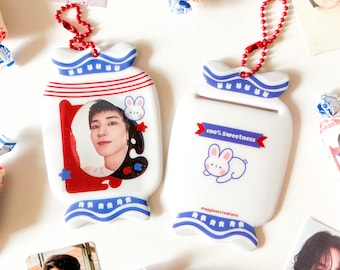 May include: Two white ceramic candy-shaped keychains with blue and red accents. One keychain features a bunny and the text "100% sweetness" and the other keychain has a photo frame with a picture of a person. Both keychains have a red chain attached.