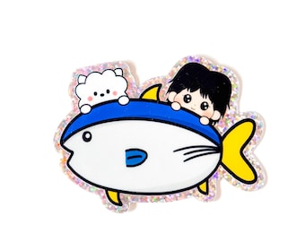 May include: A whimsical sticker featuring a cartoon fish in blue and white, with yellow fins. Two cartoon characters, a boy and a fluffy white animal, are peeking over the fish. The sticker has a glittery, iridescent border.
