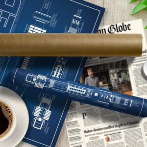 May include: A blue and white blueprint style poster with a diagram of a lightsaber, titled "Ezra Bridger's Lightsaber". The poster is rolled up and sitting on a newspaper with a coffee cup and saucer.
