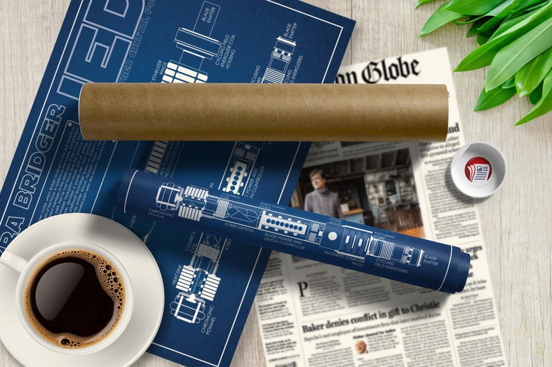 May include: A blue and white blueprint style poster with a diagram of a lightsaber, titled "Ezra Bridger's Lightsaber". The poster is rolled up and sitting on a newspaper with a coffee cup and saucer.