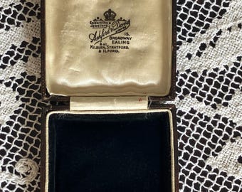 May include: An open, vintage jewelry box with a dark brown exterior and a black velvet interior. The inside lid is lined with cream-colored satin and features the text "Goldsmiths & Jewellers Ashford & Davis Limited" with addresses in Ealing, Kilburn, Stratford, and Ilford.