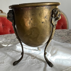 Peut inclure: Un pot en laiton à trois pieds et des poignées en tête de lion. Le pot est sur une nappe blanche.