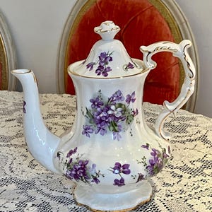 Puede incluir: Tetera de porcelana blanca con tapa, adornada con motivos florales morados y ribetes dorados. La tetera presenta un pico curvo y un asa decorativa. La base tiene un borde festoneado. Una pieza clásica para servir té.