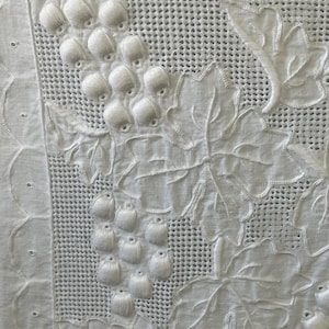 Pode incluir: Toalha de mesa de algodão branco com um delicado desenho bordado de uvas e folhas. O bordado é em relevo e texturizado, criando um efeito tridimensional.