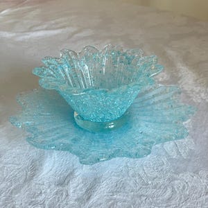 May include: A light blue glass bowl and plate set, shaped like a flower. The bowl has a textured finish and sits on a clear, circular base. The plate has a scalloped edge. The set is displayed on a white, patterned tablecloth.