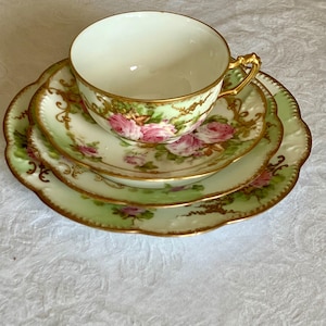 Può includere: Un servizio da tè vintage con una tazza, un piattino e un piattino da dessert. Il set ha uno sfondo verde chiaro con disegni floreali di rose rosa e finiture dorate. La tazza ha un manico dorato e i bordi dei piatti sono smerlati.