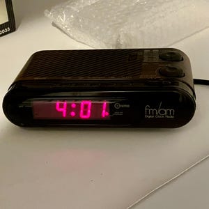 May include: Black Cosmo digital clock radio showing 4:01 in pink. The clock has an FM/AM radio function and two control knobs. The rectangular clock has a textured top. The time is displayed in a digital format.