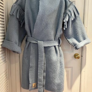 May include: A light blue coat with fringe detail on the shoulders and sleeves. The coat has a matching belt and black stitching accents. A small tag is sewn on the lower left side. The coat is hanging on a white door.
