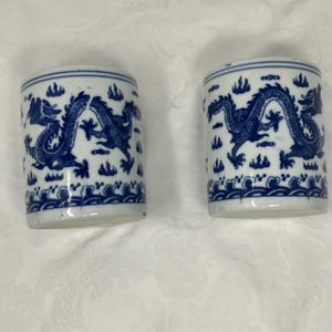 May include: Two cylindrical ceramic cups with a blue and white dragon design. The cups feature a blue dragon motif with cloud and wave patterns. The cups are on a white surface.