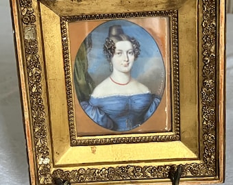 May include: A framed miniature portrait of a woman in a blue dress and red necklace. The portrait is set in an ornate gold frame with a decorative floral pattern. The frame is supported by a small, dark metal stand.
