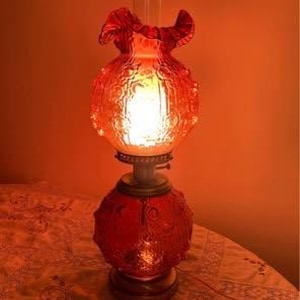 Può includere: Una lampada vintage in vetro rosso con un motivo strutturato. La lampada ha una base rotonda, una sezione centrale bulbosa e una parte superiore a balze. La lampada è illuminata, emettendo un bagliore arancione caldo. La lampada è su una superficie a motivi.