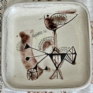 Puede incluir: Un plato cuadrado de cerámica blanca con un diseño abstracto marrón y negro que representa un pájaro. El plato está firmado "Espinosa 76".
