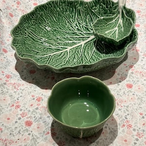 May include: A green ceramic serving platter and a small bowl. The platter is shaped like a large leaf with a smaller leaf-shaped dish attached. The bowl is round with a leaf-like design. The set is on a floral patterned surface.