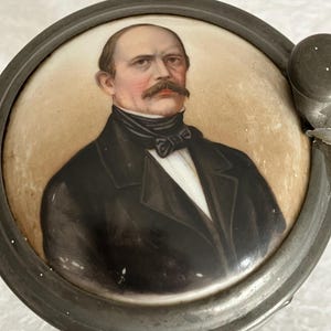 May include: An antique ceramic lid with a portrait of a man with a large moustache, wearing a black coat and bow tie. The portrait is set within a metal frame, showcasing a vintage aesthetic.