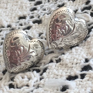 以下が含まれることがあります： 彫刻された花の模様が施された、銀色のハート型のイヤリング2つ。 イヤリングは詳細なボーダーと研磨された仕上げが特徴です。 背景は白い、オープン織りの生地です。