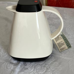 May include: A white insulated carafe with a black base and lid, featuring a curved handle. The lid has a red button. A tag with the word "DESIGN" is attached to the carafe.
