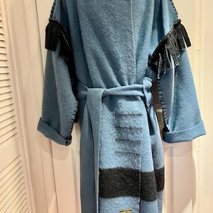 Op de afbeelding: Een lichtblauwe jas met capuchon, riem en zwarte franjes op de mouwen. De jas heeft een zwart streepontwerp aan de voorkant en een kleine patch aan de onderkant.