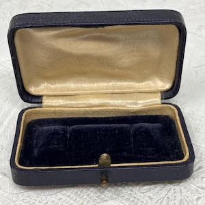 May include: Open navy blue jewellery box with a gold satin interior and a dark blue velvet insert. The box is rectangular with a hinged lid and a brass clasp. The exterior has a textured finish.