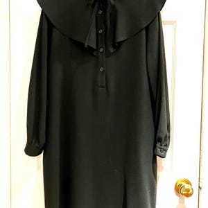 Könnte beinhalten: Ein schwarzes Kleid mit einem großen, abgerundeten Kragen und langen Ärmeln. Das Kleid hat eine Knopfleiste vorne und eine Krawatte am Kragen. Das Kleid hängt an einem Kleiderbügel vor einer weißen Tür.