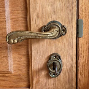 Puede incluir: Primer plano de una puerta de madera con un tirador y una cerradura de latón antiguo. El tirador tiene un diseño ornamentado y curvado. La puerta es de madera clara con veta visible.