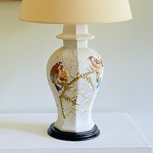 May include: A white ceramic table lamp with a beige lampshade. The lamp base features a painted design of birds perched on branches with flowers. The base has a black wooden base.