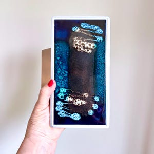 May include: Rectangular abstract art piece with a white frame. The artwork features a dark, vertical central shape with white and brown accents, surrounded by blue and teal patterns. The piece is held by a hand with red nail polish.