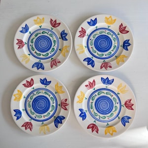 May include: Four white ceramic plates with a floral design. Each plate features a blue spiral center, surrounded by a green and blue border, and alternating red, yellow, and blue tulip-shaped flowers around the rim. A decorative tableware set.