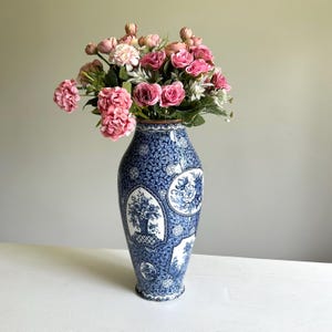 Könnte beinhalten: Eine hohe Keramikvase mit einem komplizierten blau-weißen geometrischen Muster und ovalen Feldern, die florale Illustrationen enthalten. Die Vase ist mit einem großen Strauß aus künstlichen rosa Rosen und kleinen weißen Blumen gefüllt.