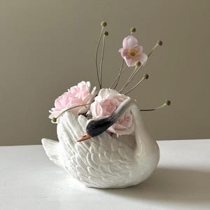 Könnte beinhalten: Ein weißer, schwanenförmiger Übertopf aus Keramik mit glänzender Oberfläche, schwarzem Hals und orangefarbenem Schnabel. Der Schwan ist mit mehreren hellrosa Rosenblüten und dünnen, hohen Stielen mit kleinen, runden, grünen Samenkapseln gefüllt.
