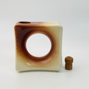 May include: A square ceramic vase with a large circular opening, featuring a gradient of brown and beige tones. A small, decorative wooden stopper with a geometric pattern is next to the vase.