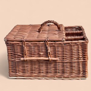 May include: A brown wicker picnic basket with a hinged lid and a woven handle. The basket has a rectangular shape with a compartment for utensils. The basket is made of woven brown wicker.