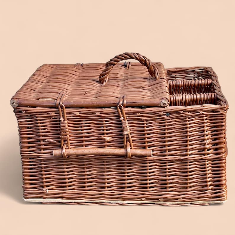 May include: A brown wicker picnic basket with a hinged lid and a woven handle. The basket has a rectangular shape with a compartment for utensils. The basket is made of woven brown wicker.