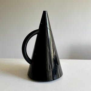 Könnte beinhalten: Ein glänzend schwarzer konischer Krug mit einer glatten, reflektierenden Oberfläche und einem geschwungenen Griff. Das Gefäß hat eine breite kreisförmige Basis, die sich nach oben zu einer Spitze verjüngt und eine markante geometrische Form bildet.