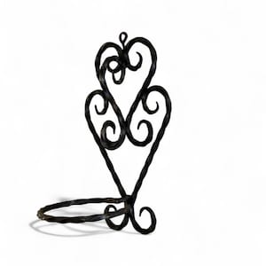 Könnte beinhalten: Schwarzer Kerzenhalter aus Schmiedeeisen mit herzförmigem Design. Der Halter hat einen kreisförmigen Sockel und eine dekorative Oberseite mit verschlungenen Herzen und Locken. Das Metall hat eine strukturierte Oberfläche vor weißem Hintergrund.