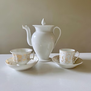 Puede incluir: Un juego de café de porcelana blanca con una cafetera grande y dos tazas con platillos. El juego tiene detalles florales dorados. La cafetera mide aproximadamente 25 cm de alto. Las tazas y platillos miden aproximadamente 5 cm de alto.