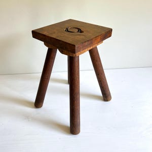 May include: A small, three-legged wooden stool. The square seat has a circular indentation. The stool is made of dark brown wood. The legs are rounded and taper towards the bottom. The stool's height is approximately 30 cm.