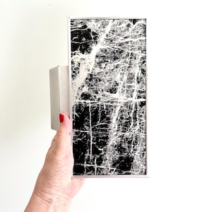 Può includere: Applique da parete rettangolare con un design nero e bianco simile al marmo. L'applique ha una cornice argentata e una piccola base rettangolare. Il design presenta una rete di venature bianche su sfondo nero.