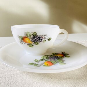 May include: A white teacup and saucer set. The cup and saucer feature a fruit design with pears, grapes, and green leaves. The cup has a handle and sits on a matching saucer. The set is made of opaque white glass.