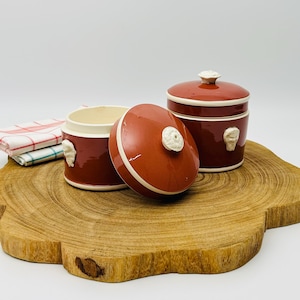 Peut inclure: Deux pots en céramique avec couvercles, dont un ouvert, de couleur marron foncé avec des accents blancs. Les couvercles ont un bouton blanc. Les pots ont un intérieur crème et sont décorés d'un motif en relief blanc. Les pots sont sur une surface en bois.
